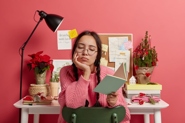 Free photo serious asian student learns information from textbook, does school homework, wears round glasses and sweater, poses against desktop