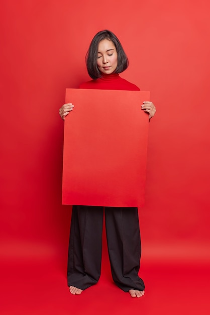 Serious Asian woman holds red advertising board recommends to place your information here displays banner weras stylish outfit poses against bright studio wall