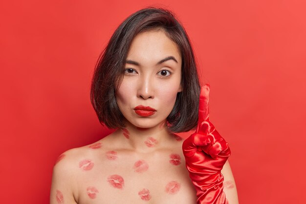 Serious attentive Asian woman raises index finger wears gloves stands shirtless with lips traces wears red lipstick poses indoor has excellent idea