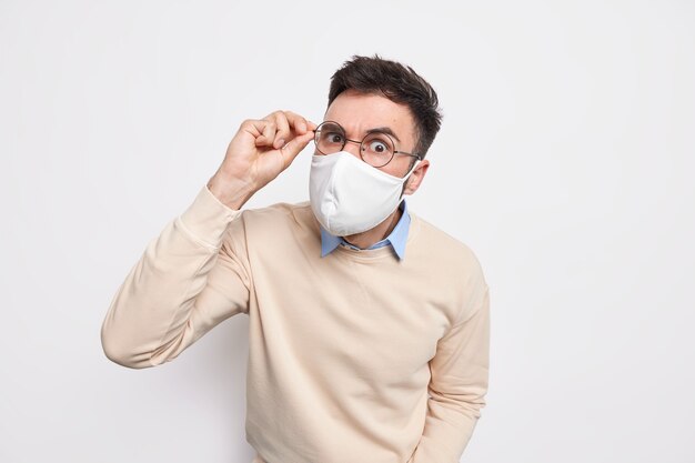 Serious attentive brunet man wears protective mask in public place keeps hand on spectacles prevents himself from coronavirus dressed casually 