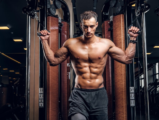 Serious attractive man is doing exercises with training apparatus at dark gym club.