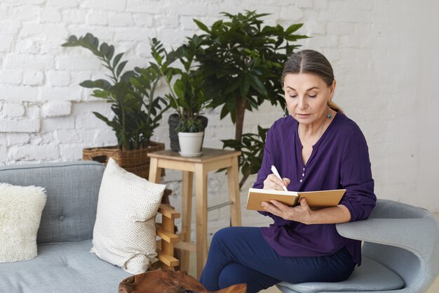 Serious attractive mature female business coach having focused concentrated look while writing down in her notebook, fixing appointment with client, sitting on chair in modern apartment interior