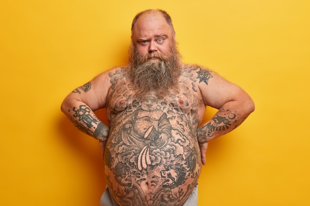 Free photo serious bearded fat man has thick beard, tattooed body and big belly, looks from under eyebrows, keeps hands on waist, isolated on yellow wall. obesity, liposuction, weigh loss concept