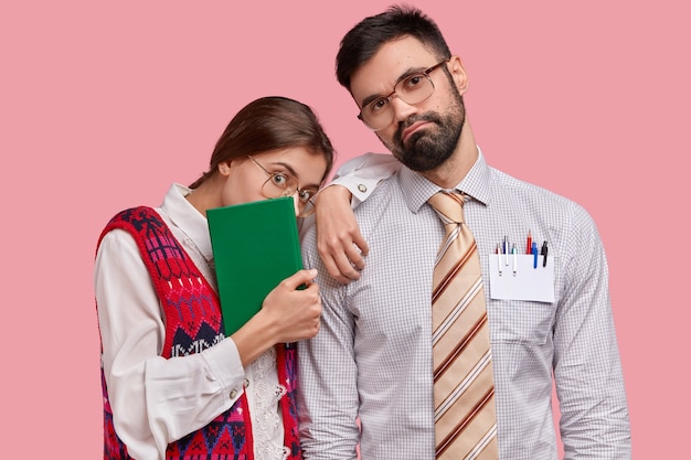 Serious bearded man in formal clothes tilts head, beautiful female wonk carries green textbook, wears old style clothes, leans at shoulder of partner