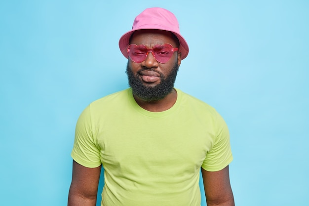 Serious bearded man looks directly at front wears trendy sunglasses hat and casual green t shirt isolated over blue wall