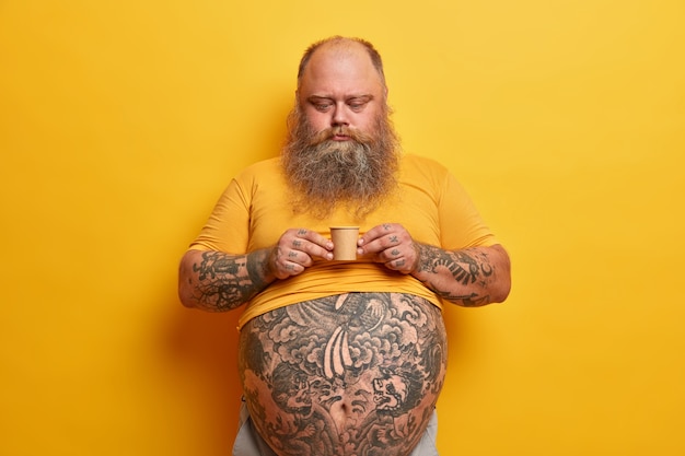 Serious bearded man with big tummy, tattooed arms and belly, holds very small carton cup of coffee containing much sugar, enjoys caffeine aromatic beverage, wears yellow t shirt, poses indoor alone