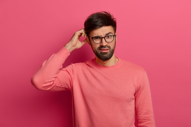 Free photo serious bearded young man scratches head, thinks deeply about how to solve problem