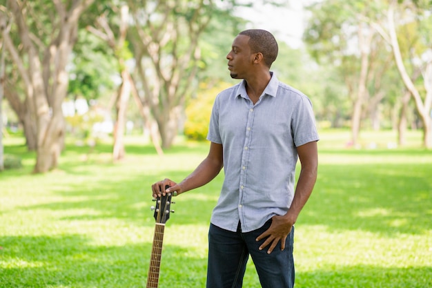 Serious black man holding guitar by headstock in park