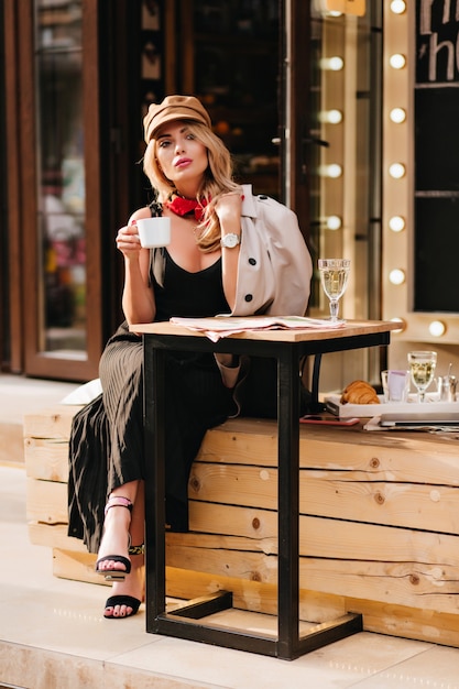 Free Photo serious blonde girl in black shoes chilling in outdoor cafe and enjoying tea. attractive young woman wears stylish sandals and brown hat looking away, holding cup of coffee.