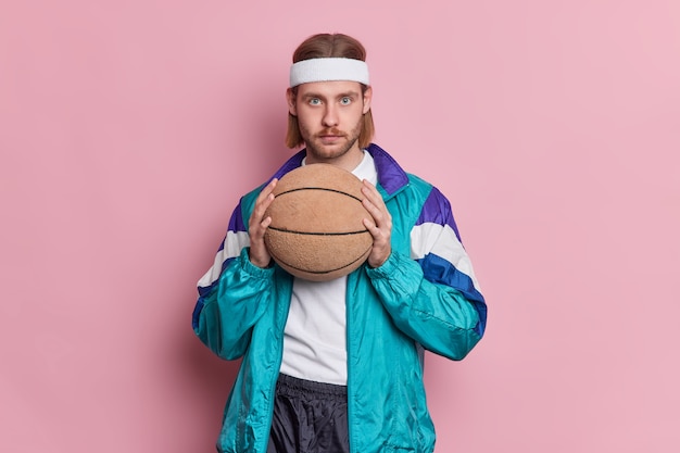 Serious blue eyed male basketball player with stubble long hair holds ball ready for playing game wears white headband and sportsclothes.