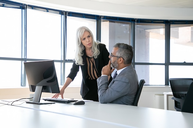 Serious boss and manager discussing report, talking while sitting and standing at workplace with panoramic window . Business communication concept