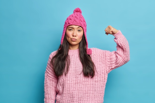 Free photo serious brunette woman raises arm and shows muscles being self confident and full of power wears knitted jumper pink hat feels strong healthy isolated over blue wall