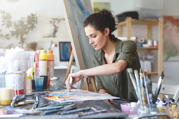 Free photo serious brunette young beautiful woman sitting in art studio, taking colorful paints from tube while creating great masterpiece on easel, being preoccupied with her work, having nice imagination