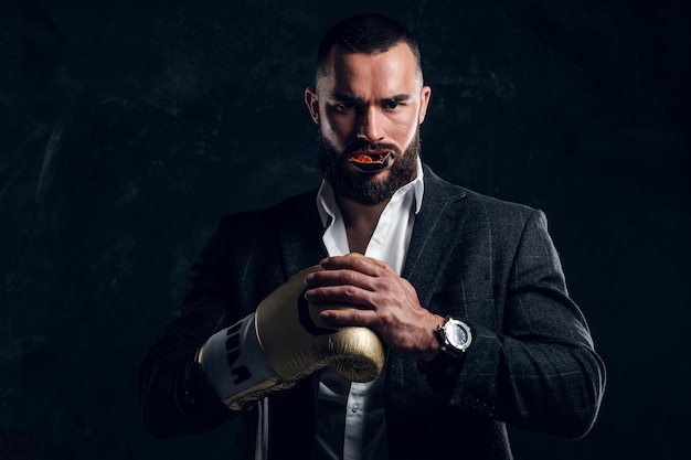 Serious brutal man in suit and golden boxing glove is posing for photographer at dark photo studio.