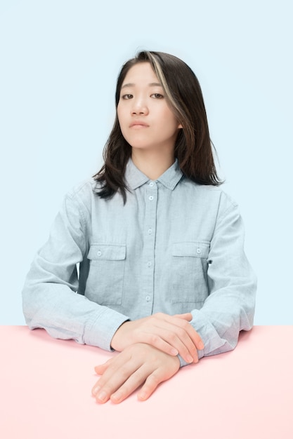 Serious business woman sitting at table, looking at camera isolated on trendy blue studio background. Beautiful, young face.