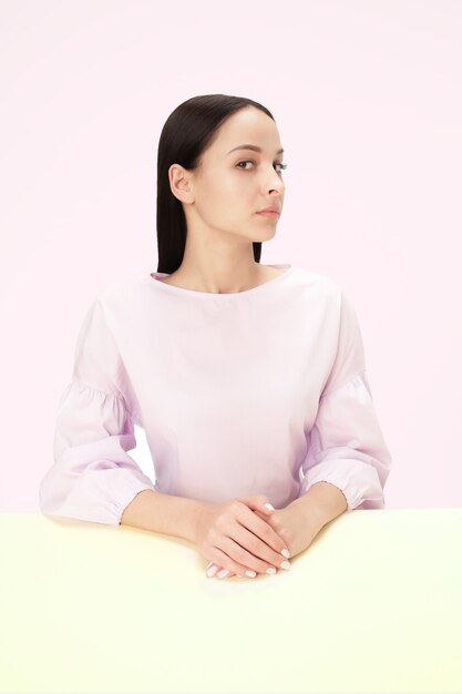 Serious business woman sitting at table on a pink studio background. The portrait in minimalism style