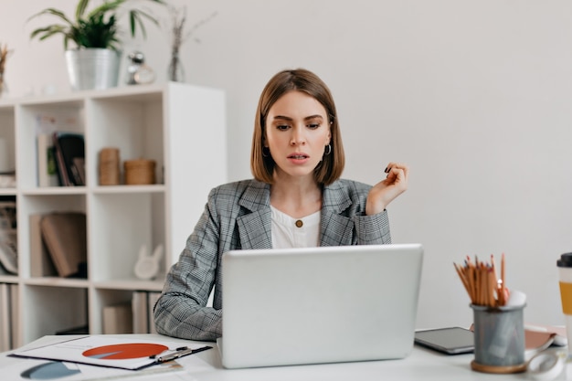 Serious business woman with anxiety looks in laptop. Portrait of girl with short haircut in white office.