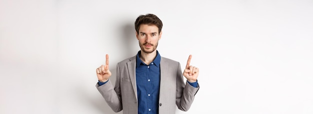 Free Photo serious businessman with beard wearing suit pointing fingers up look here gesture advertising on whi