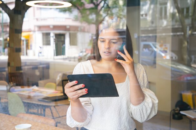 Serious businesswoman talking on cell, using tablet and looking at screen