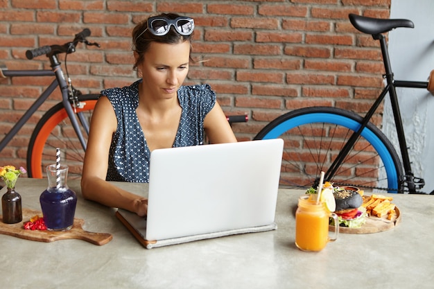 Serious businesswoman with shades on her head checking email on her modern laptop computer during lunch on weekend. Self-employed female using notebook pc for remote work