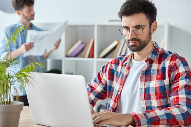 Serious bussinessman with thick beard, analyzes income charts and graphs on laptop computer, dressed in checkered shirt