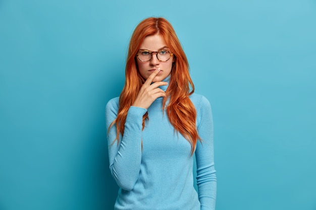 Free photo serious calm woman has natural long ginger hair keeps hands on lips and looks with concentrated thoughtful expression.