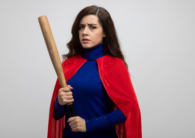 Serious caucasian superhero girl with red cape holding baseball bat isolated on white wall with copy space