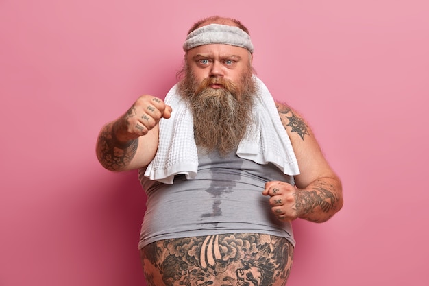 Serious chubby bearded man shows clenched fists, suffers from extra weight, goes in for sport, has sweaty body and tattooed arms, poses against pink wall. Slimming and dieting concept.