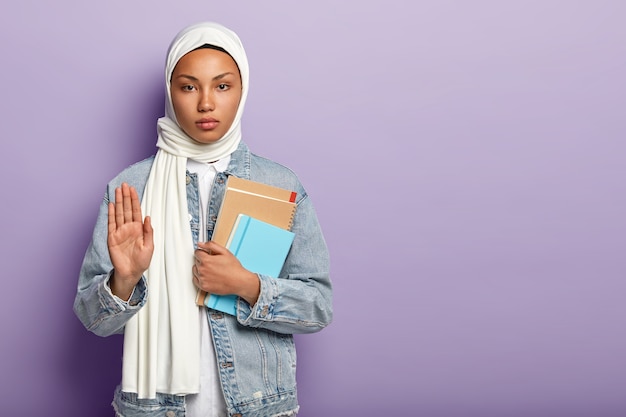 Serious confident Muslim woman holds notepads, shows palm as sign of refusal or rejection, wears white scarf and denim coat, asks to wait minute, poses over purple wall, blank space