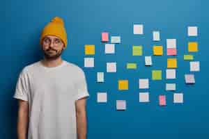 Free photo serious contemplative man with beard, dressed casually, thinks about writing diploma, uses adhesive notes to write down information to remember.