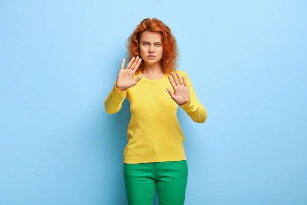 Free photo serious discontent woman has wavy red hair, shows stop gesture, keeps palms outstretched at camera, refuses something