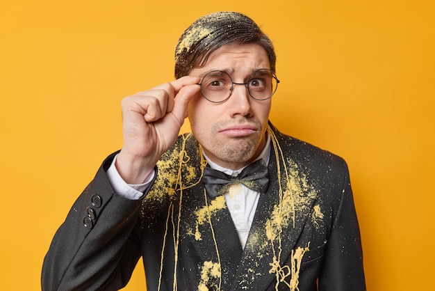 Free photo serious displeased man keeps hand on rim of spectacles focused attentively at camera doesnt like something dressed in festive formal suit smeared with serpentine spray isolated over yellow wall