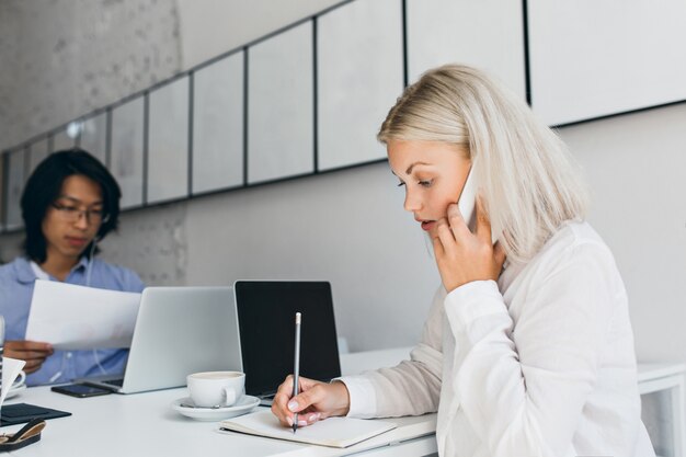 Serious fair-haired woman talking on phone and writing something on paper