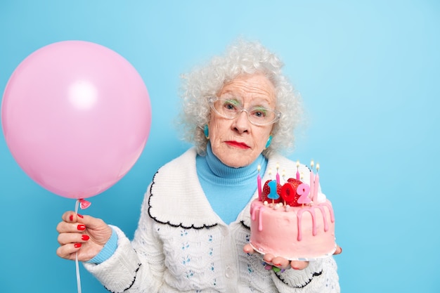 Serious fashionable elderly lady looks directly, celebrates birthday poses with festive cake and inflated balloon being on pension