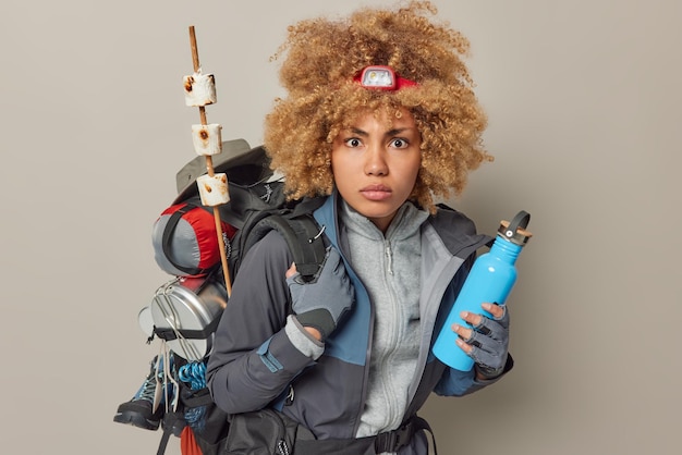 Free Photo serious female camper returns from picnic stares surprisingly carries rucksack holds bottle with fresh water dressed in sportswear isolated over grey background adventure time and recreation