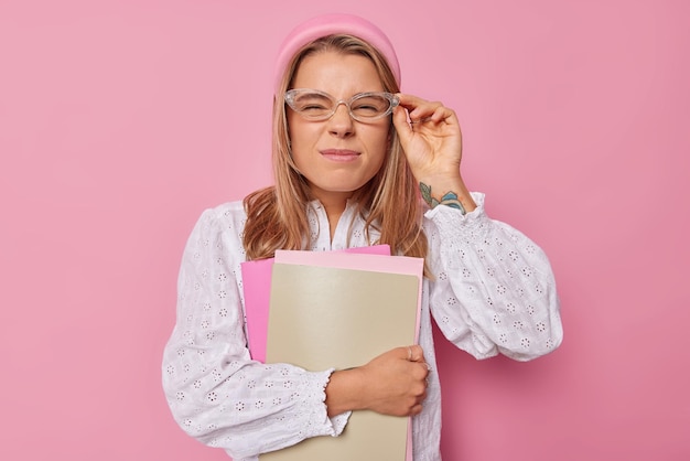 Serious female student looks through transparent spectacles dressed in white blouse poses with notebooks indoor going to have classes at university isolated over pink background. Studying concept