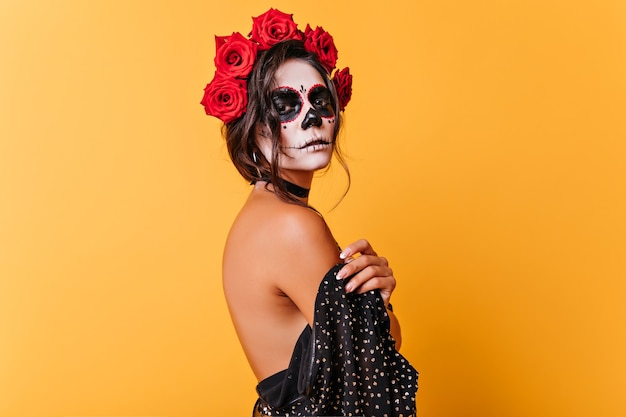 Serious girl in la muerta attire looking to camera during halloween photoshoot. Charming dead bride with roses in black hair isolated on yellow background.