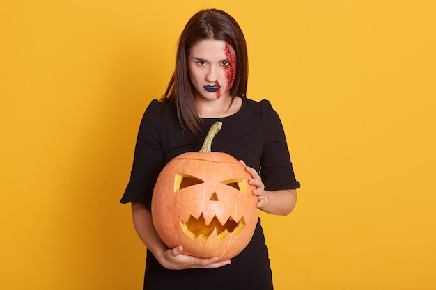 serious girl with angry facial expression standing with pumpkin in hands in studio isolated on yellow, attractive female with bloody wound on her face, halloween concept.