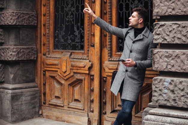 Free Photo serious good-looking man standing near old building using cell phone, and catching taxi putting finger up