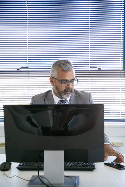 Serious grey haired businessman sitting at workplace with pc monitor and taking cellphone from desk. Front view. Communication and multitasking concept