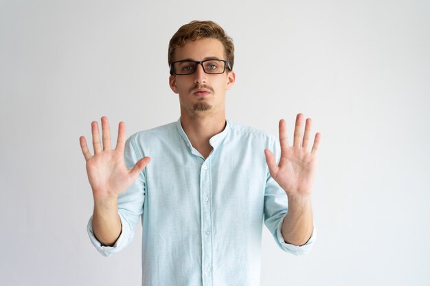 Serious handsome guy in glasses raising hands in stop sign 