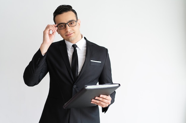 Serious handsome young businessman adjusting glasses and holding folder.