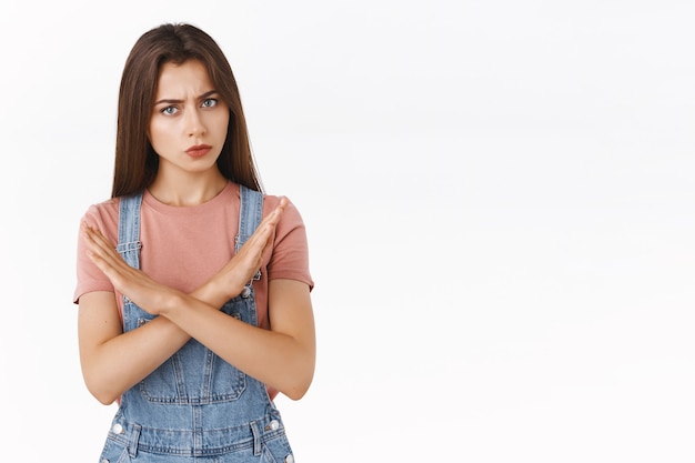 Serious-looking angry brunette girl in denim dungarees, t-shirt, making cross top stop someone, prevent bad decision, shaking head in negative reply, disagree, prohibit or refuse offer