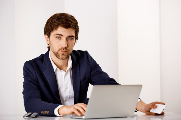 Serious-looking handsome businessman in suit, sit office desk, working on report with laptop, wait for phone call