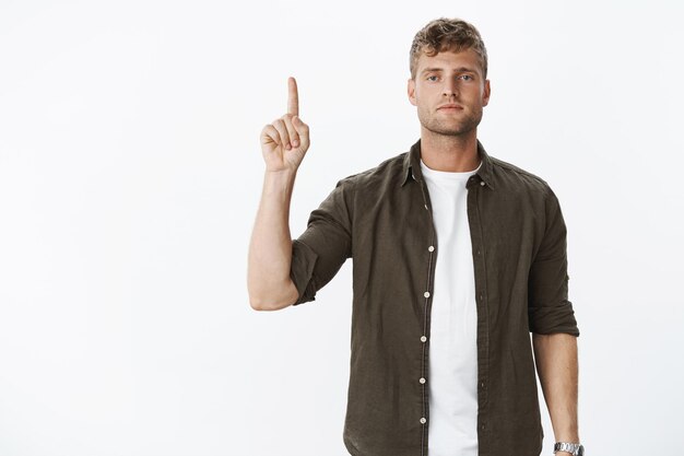 Serious-looking handsome masculine blond guy with blue eyes and bristle making poker face and pointing up with raised arm as if trying look strict and determined