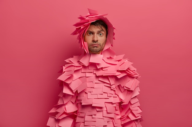 Serious looking young European man raises eyebrows, listens information attentively, prepares for office costume party, covered with adhesive notes, poses against pink wall. Monochrome