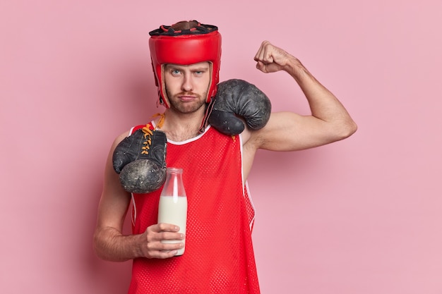 Free photo serious male boxer raises arm shows biceps drinks fresh milk to be strong wears protective hat red t shirt boxing gloves around neck demonstrates power