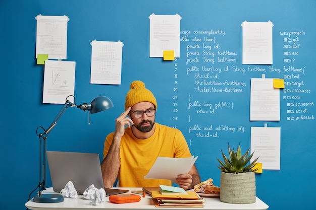 Free photo serious male employee or freelancer considers paper document, wears yellow hat and t shirt, studies on laptop computer online, works from home, looks through material, poses in coworking space