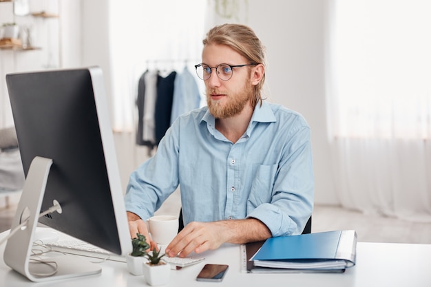 Free photo serious male trader with blonde hair, beard, wearing glasses and blue shirt, prepares financial report on company`s income, typing on keyboard of computer, sits against modern light office interior.