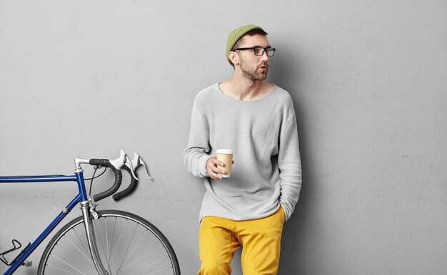 Serious male with bristle looking aside while keeping hand in pocket and holding paper cup with coffee, noticing his friend while having break after long trip alone in mountains. Lifestyle and hobby